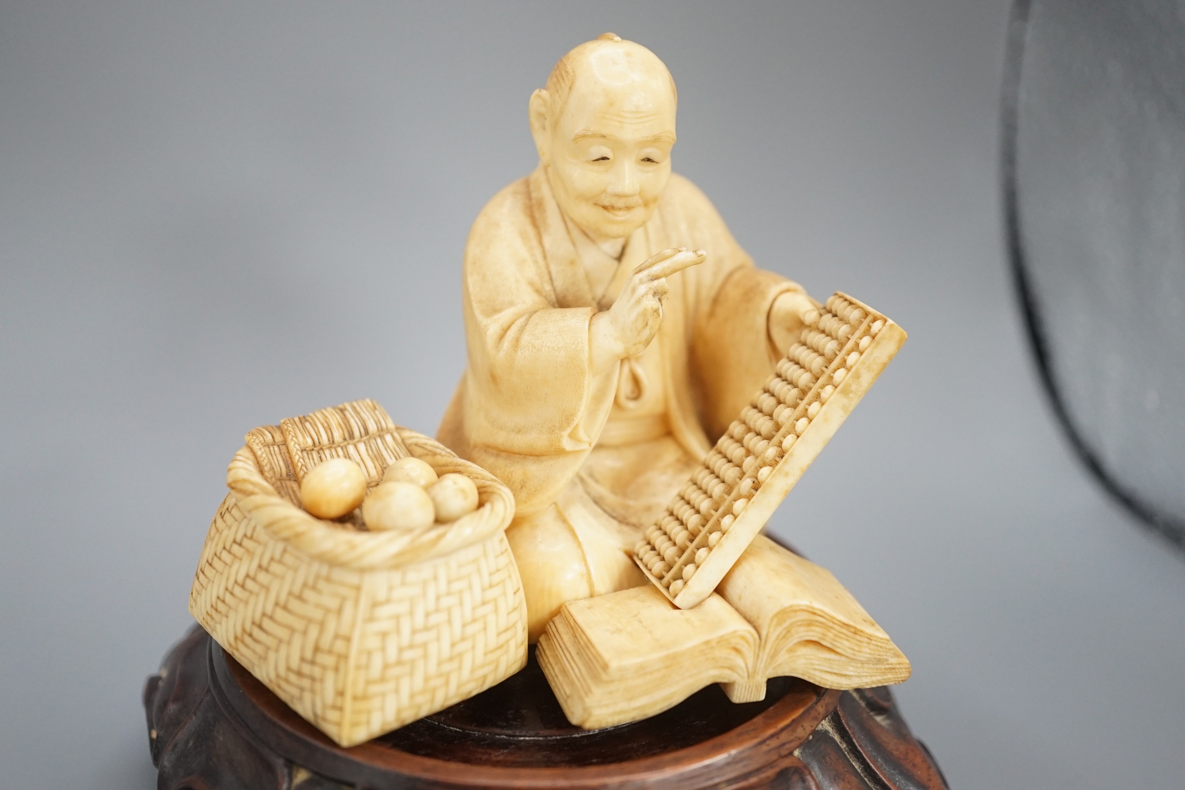 A Japanese ivory okimono of a street vendor kneeling and holding an abacus, Meiji period, 8cm, associated wood stand